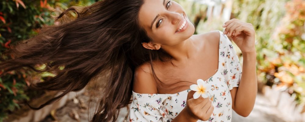 Lady with white flower in her hand plays with hair. Woman in bright summer outfit with smile posing against background of plants